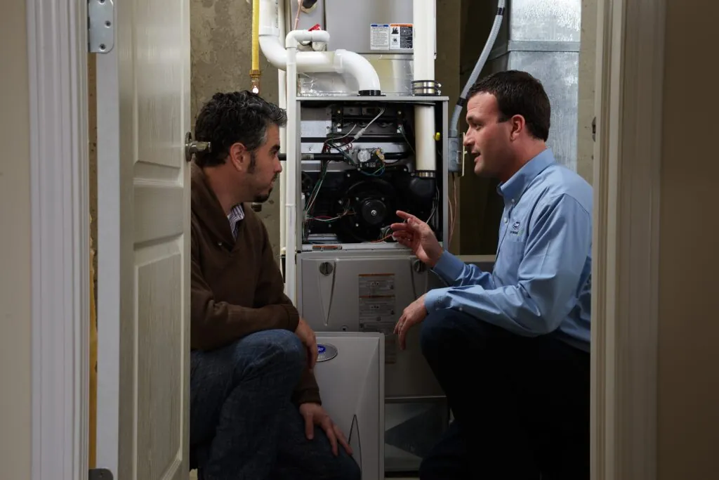 A furnace technician showing a male homeowner inside his system while both are crouched.